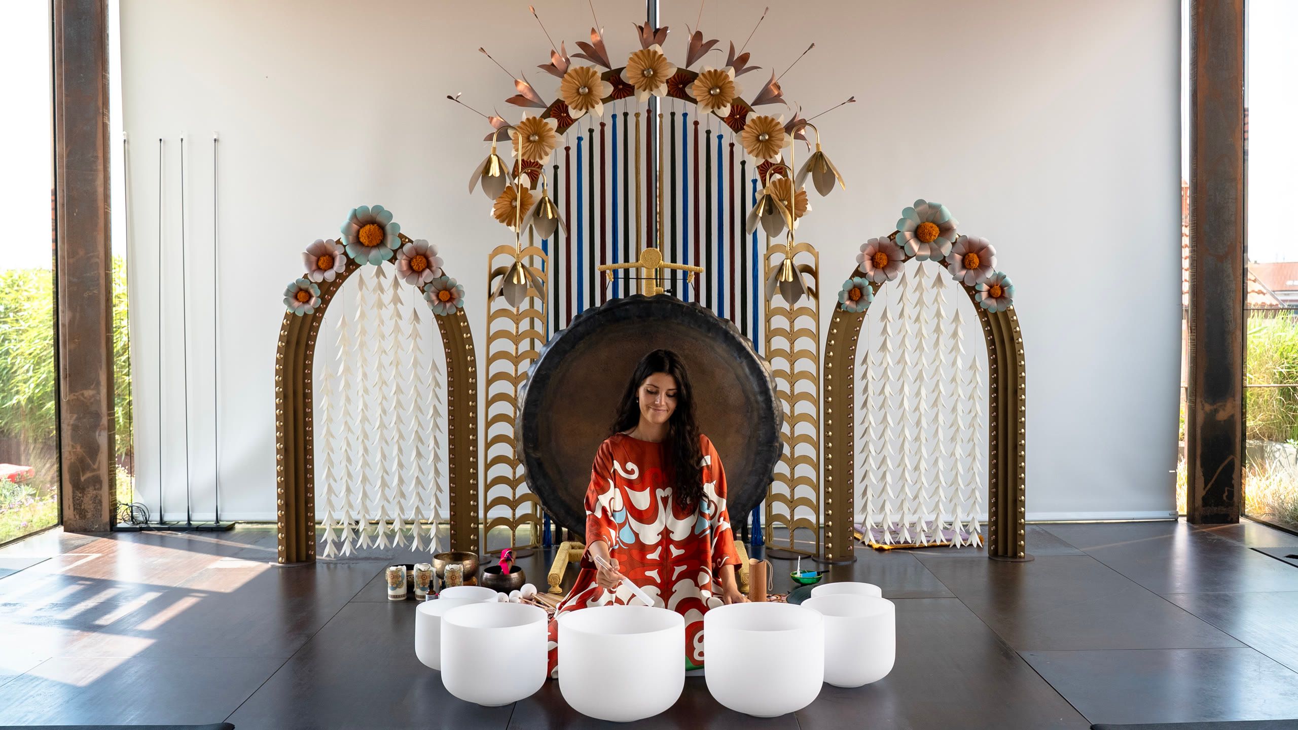 Harriet Emily playing crystal singing bowls in front of a large gong, between two arches topped with paper flowers