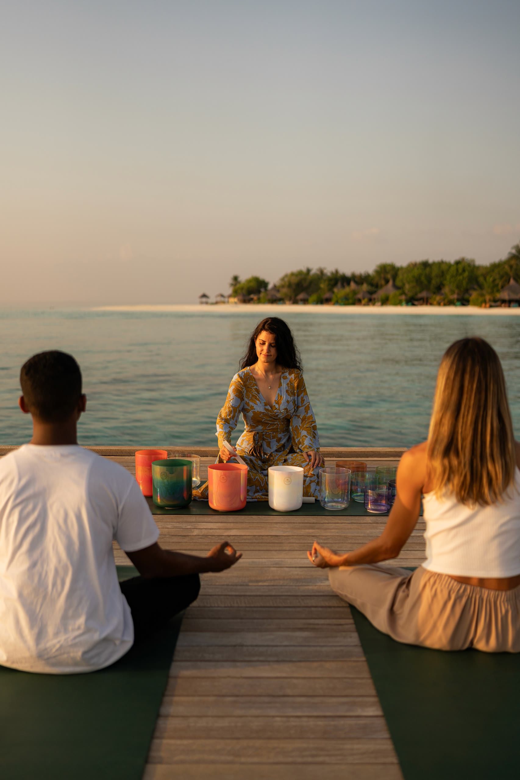 Harriet Emily playing crystal singing bowls for two guests on a seaside deck at sunrise