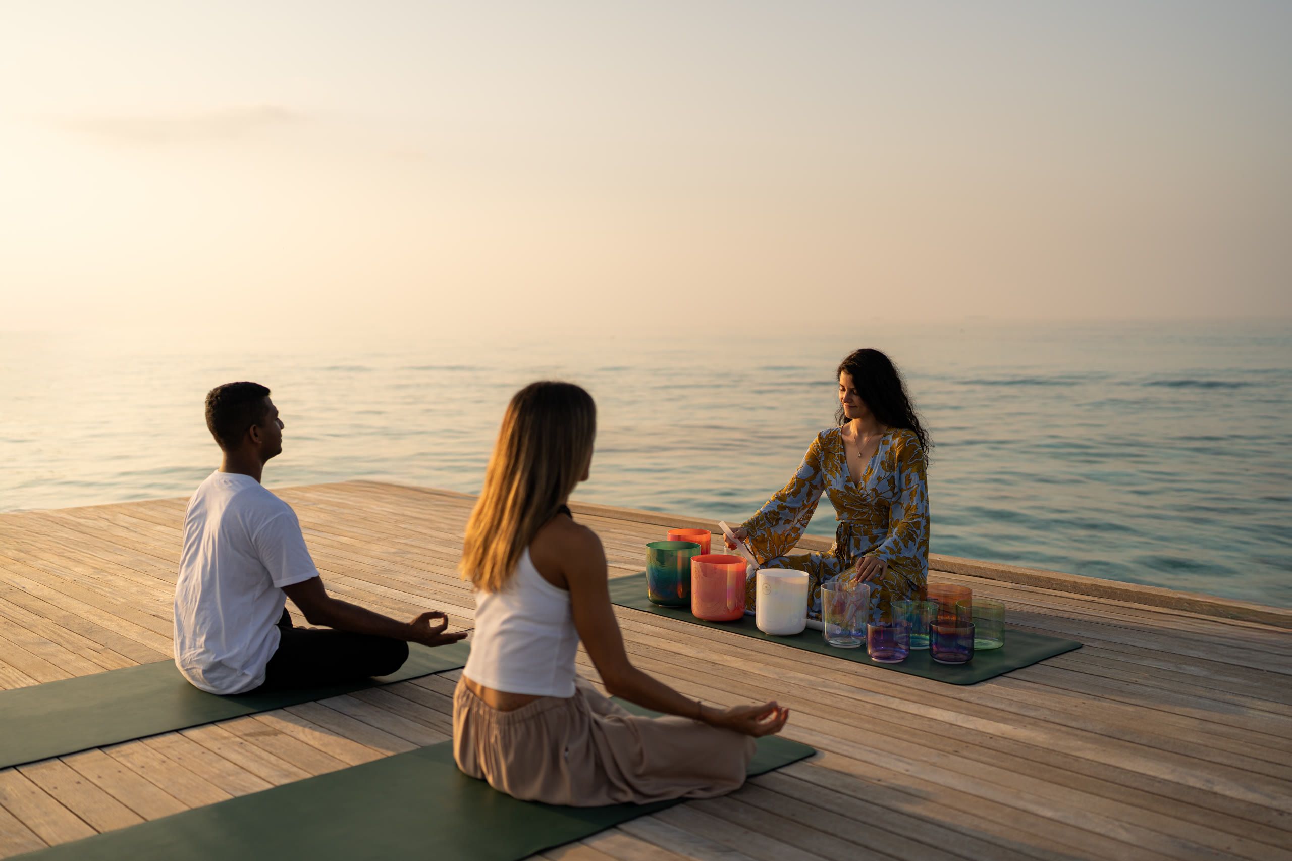 Harriet Emily playing crystal singing bowls for two guests on a seaside deck at sunrise